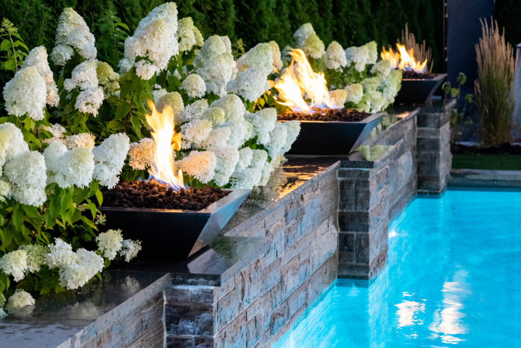 In an outdoor setting, large hydrangea bushes bloom beside a line of fire pits on a stone wall, all adjacent to a tranquil, illuminated pool.