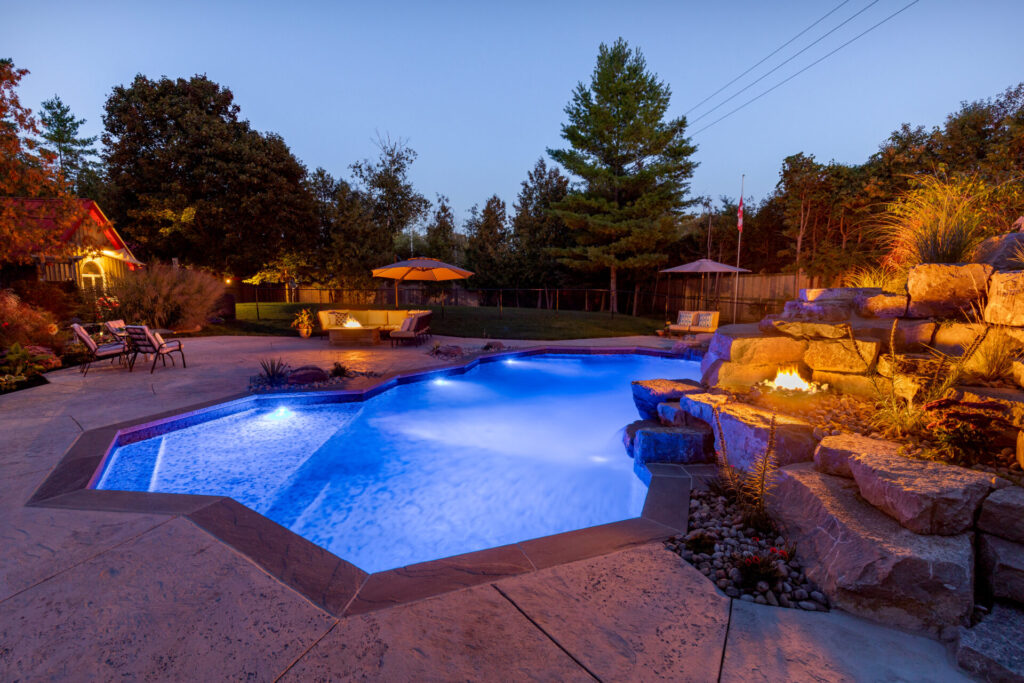A luxurious backyard with a lit pool and hot tub, surrounded by landscaping and lounge chairs, at twilight. A cozy fire feature adds ambiance.