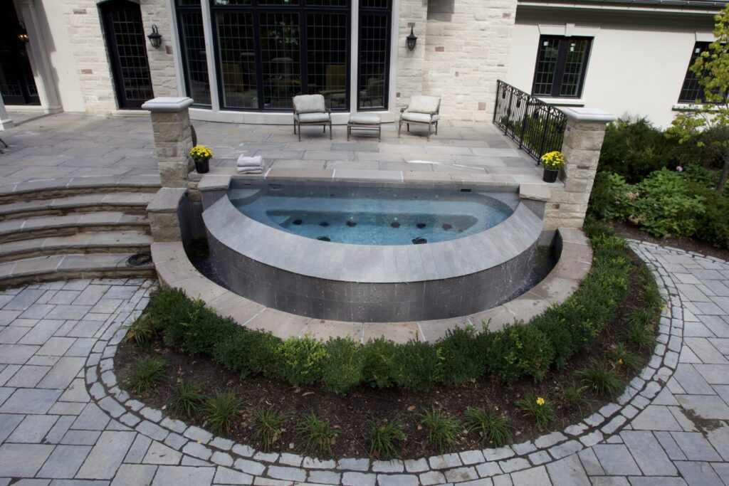 This image shows a luxurious outdoor space with a circular hot tub, stone patio, steps, landscaping, outdoor furniture, and part of a building's façade.