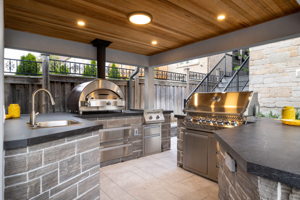 An outdoor kitchen featuring a stainless steel grill, pizza oven, sink, and storage. Covered area with wooden ceiling, and adjacent staircase leading upwards.