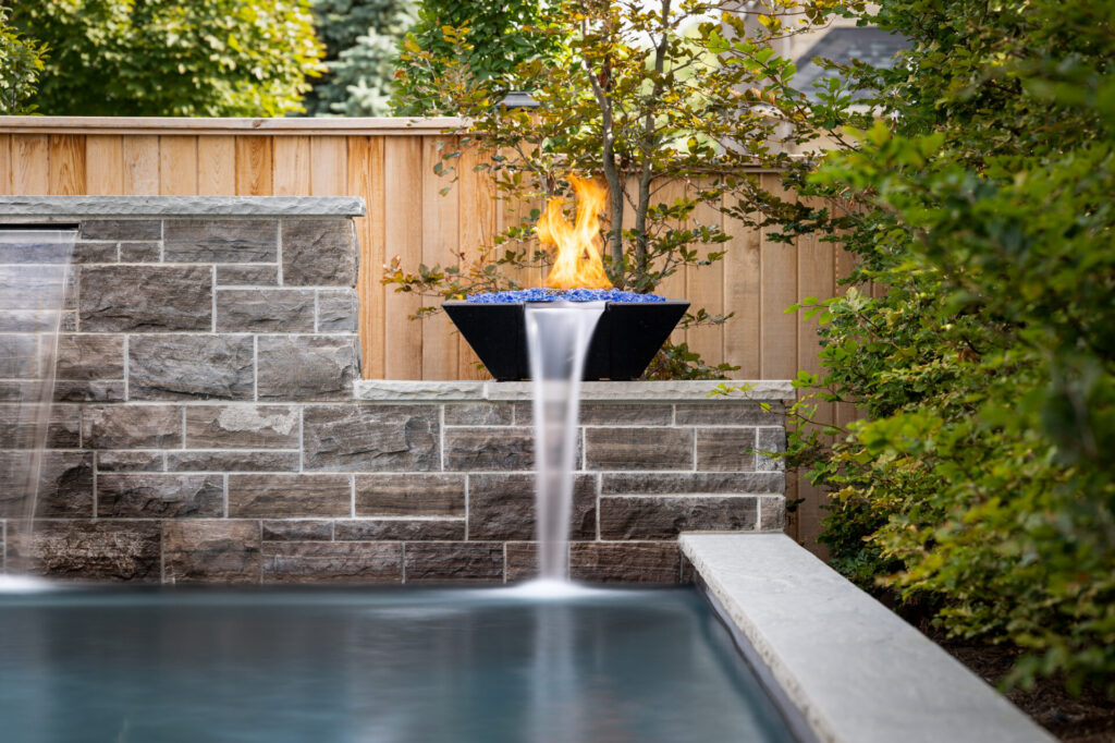 An outdoor setting features a swimming pool with a water feature, a fire pit with blue rocks and flames, surrounded by greenery and a wooden fence.