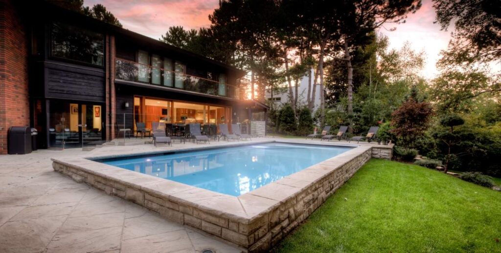 A modern two-story home with large windows overlooks a rectangular swimming pool amidst a landscaped garden with trees, at sunset.