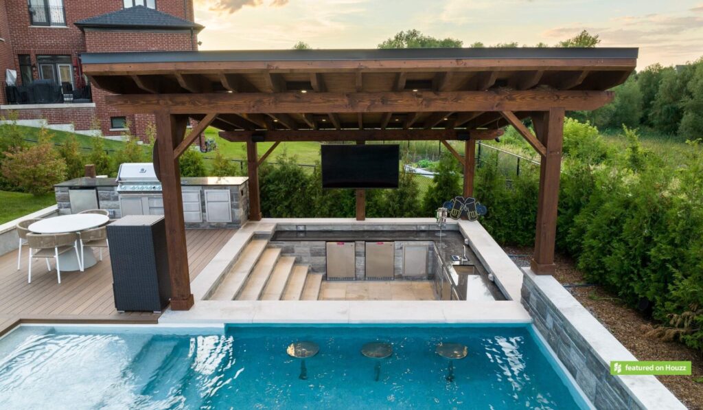 An outdoor pool with a wooden pergola, an outdoor kitchen, and a mounted TV. Lush greenery surrounds the area next to a brick house.