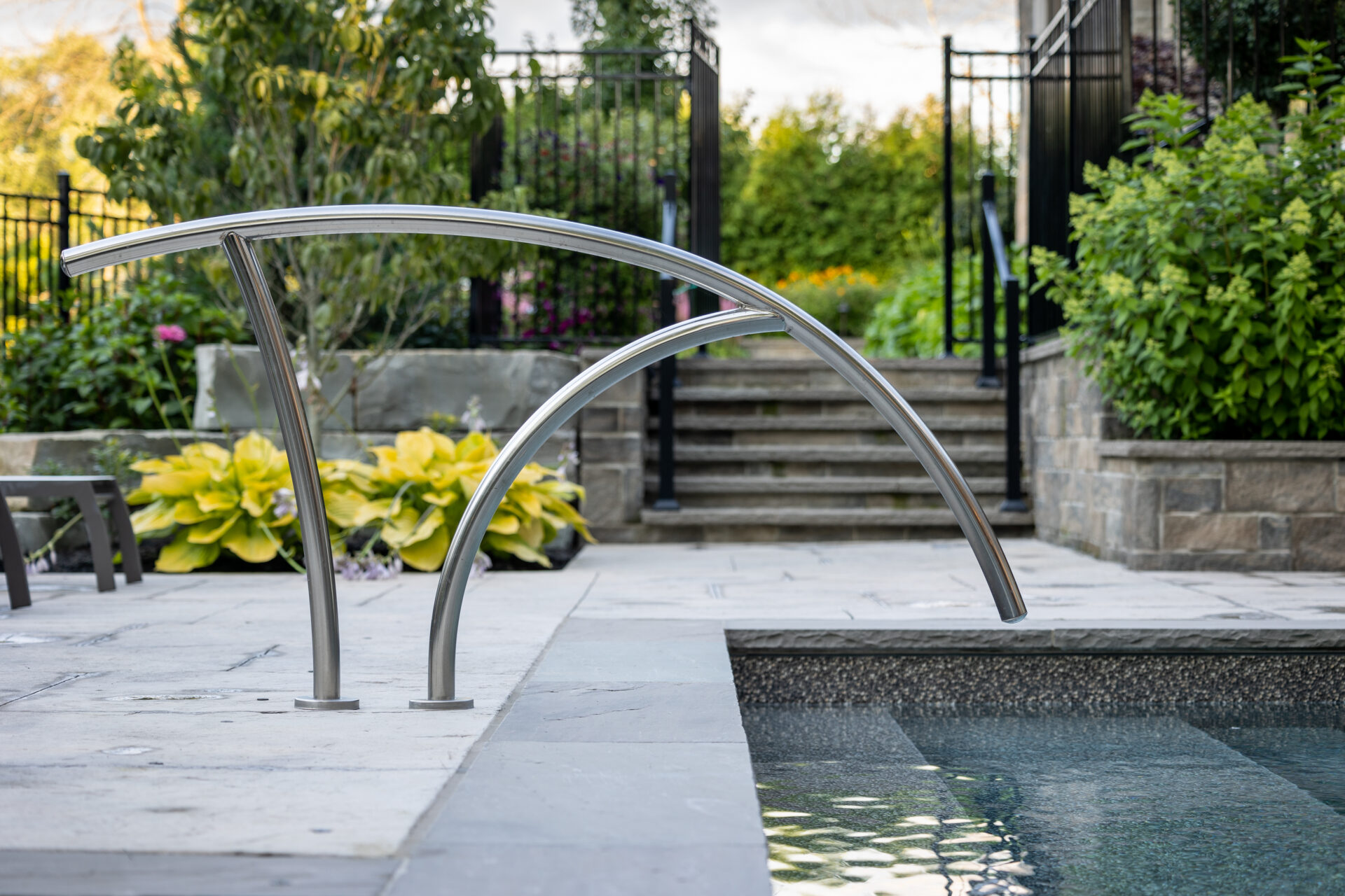 An outdoor pool with sleek, curved stainless steel handrails, surrounded by a paved area, plants, and stairs leading to a gated entrance.