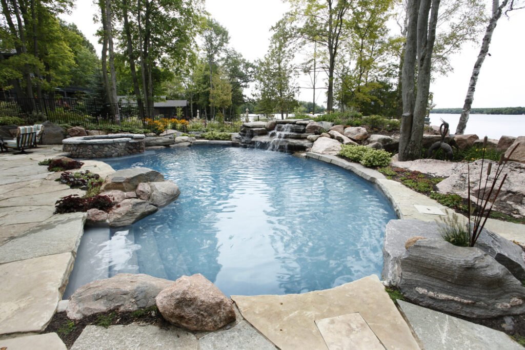An outdoor swimming pool with naturalistic rock features and a cascading waterfall, surrounded by landscaping and trees, near a body of water.