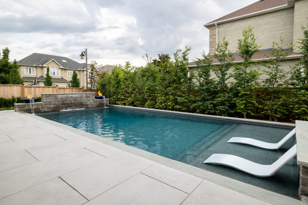 An outdoor rectangular pool flanked by lounge chairs and hedges in a backyard, with a stone fire feature and a house in the background.