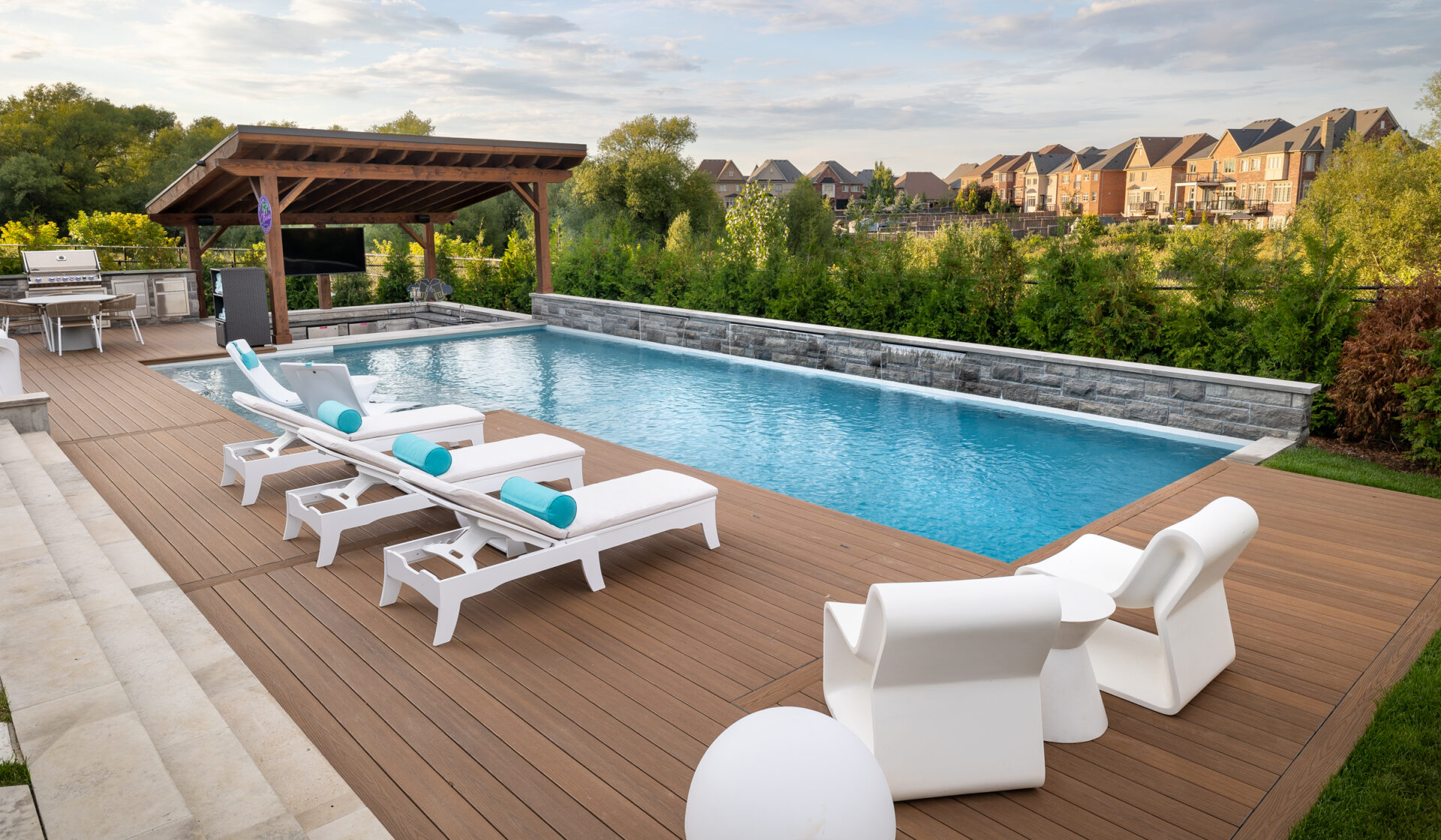 Luxurious backyard with a swimming pool, lounge chairs, outdoor kitchen, and a wooden deck overlooking a natural area and distant suburban houses.