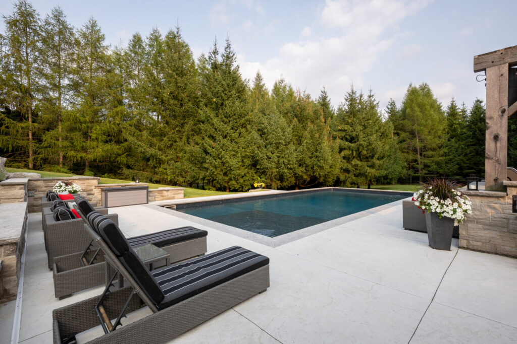 A luxurious backyard with a rectangular swimming pool, surrounded by a tiled patio and loungers, is nestled against a backdrop of tall evergreen trees.