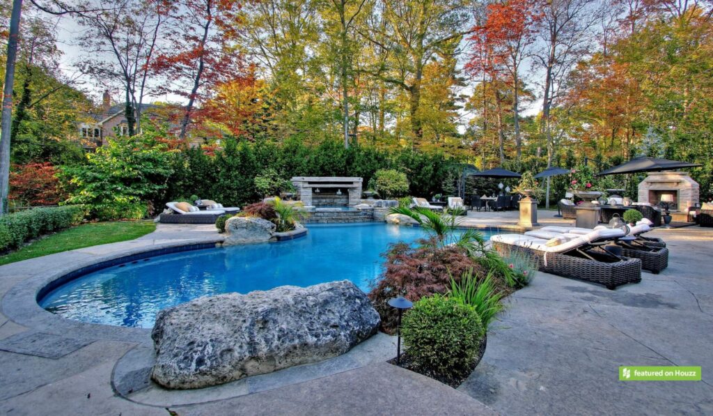 Luxurious backyard with a curved swimming pool, patio loungers, outdoor kitchen, and fireplace amidst colorful autumn trees. Elegant, inviting outdoor living space.