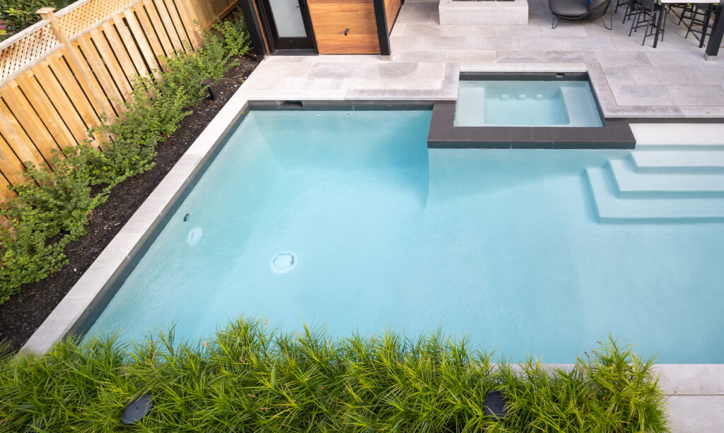 This is a sleek backyard swimming pool with an attached hot tub, surrounded by greenery, modern landscaping, and a wooden fence.