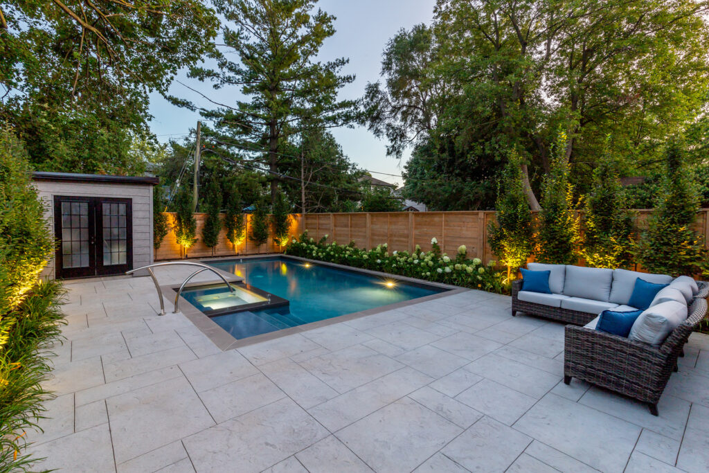 A serene backyard at dusk featuring a swimming pool, lounging area with sofa, spa, ambient lighting, and lush greenery against a wooden fence.
