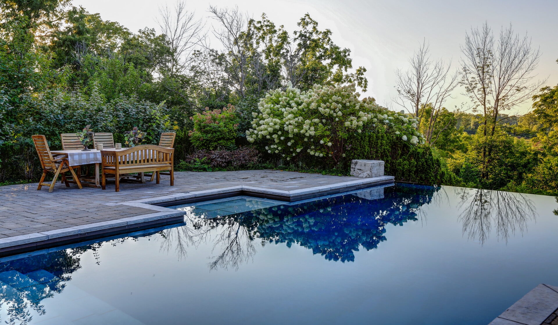 An outdoor setting with a reflective infinity pool, wooden dining furniture, surrounded by lush greenery and blooming bushes under a clear sky.