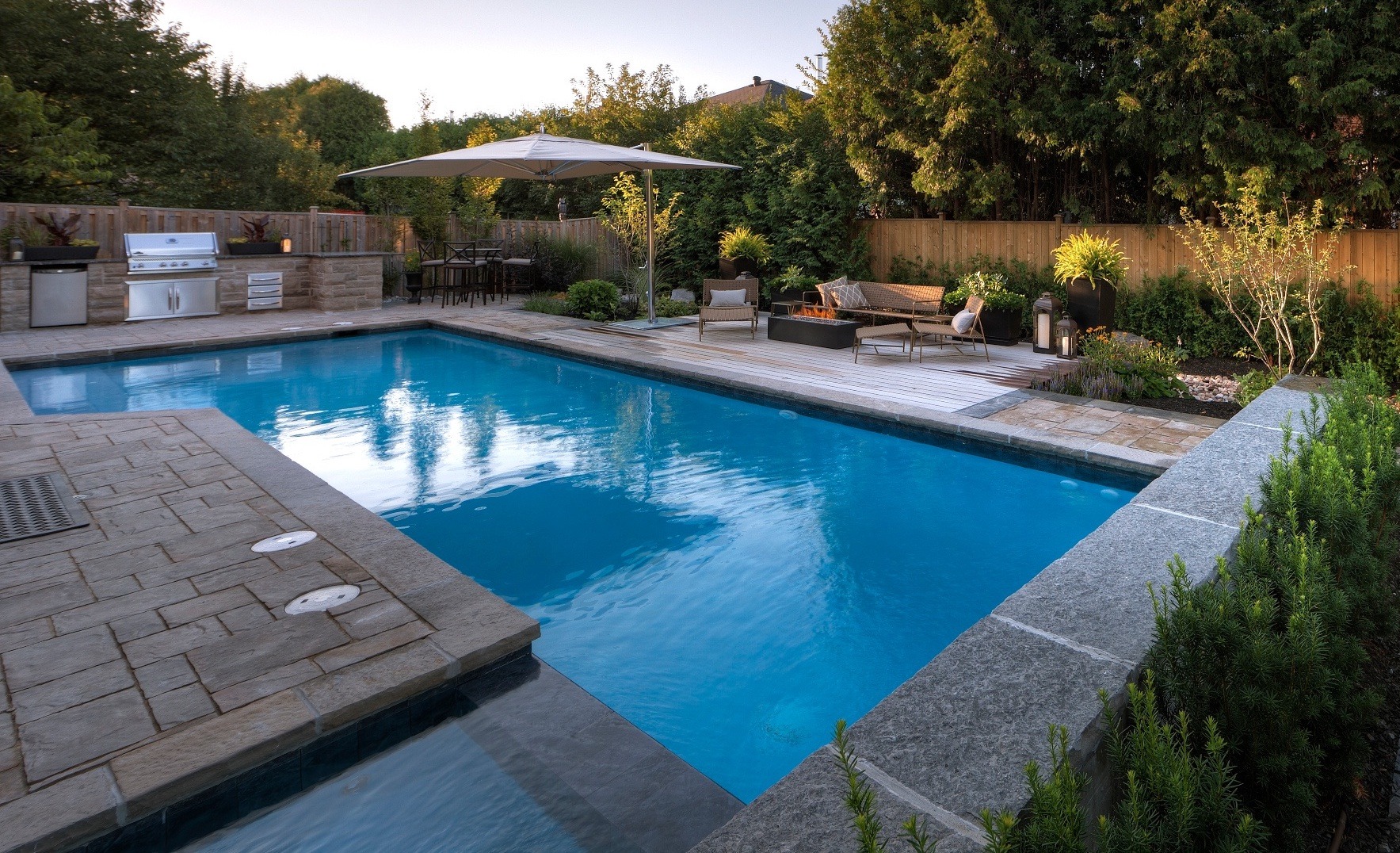 An outdoor residential setting with a clear blue swimming pool, patio with lounging chairs, an umbrella, and a grill area surrounded by trees at dusk.
