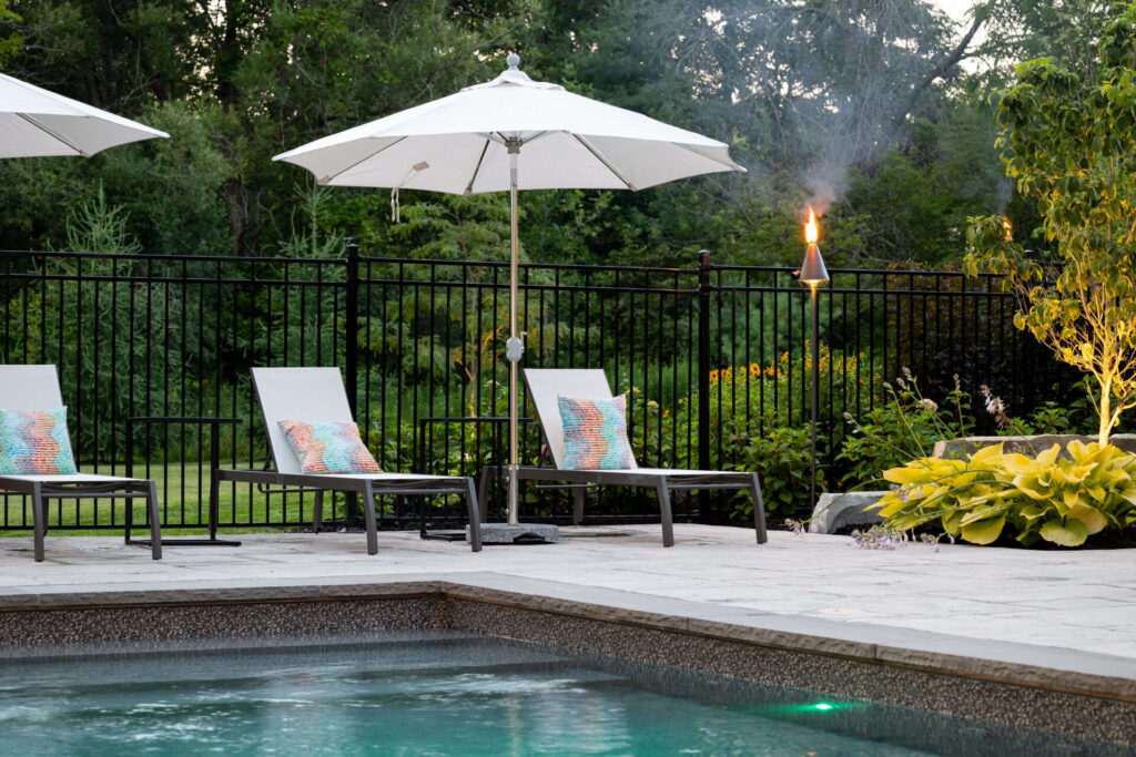 This image showcases an outdoor poolside area featuring chairs with colorful cushions under white umbrellas, a lit tiki torch, lush greenery, and a metal fence.