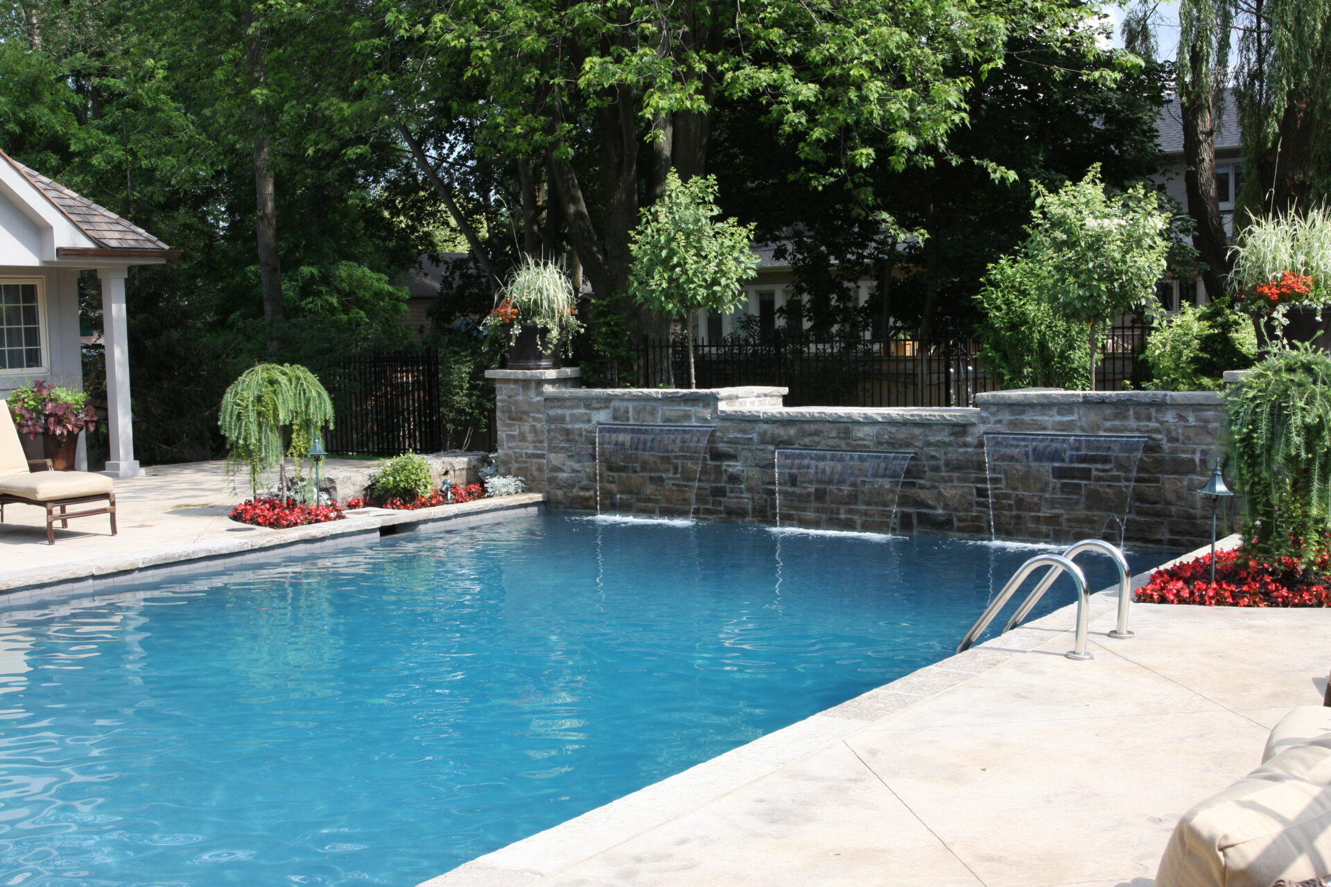 An outdoor swimming pool with a stone waterfall feature, surrounded by lounging chairs, plants, and a fence, in a landscaped backyard setting.