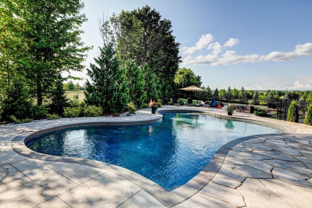 Outdoor kidney-shaped swimming pool with blue water, surrounded by stone patio, trees, a fence, lounge chairs, and clear skies overhead.