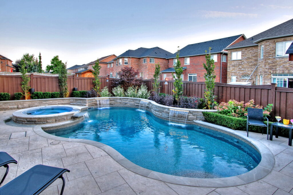 This is an image of a backyard with a curvy inground pool and hot tub, surrounded by a patio and wooden fence, with residential houses in the background.