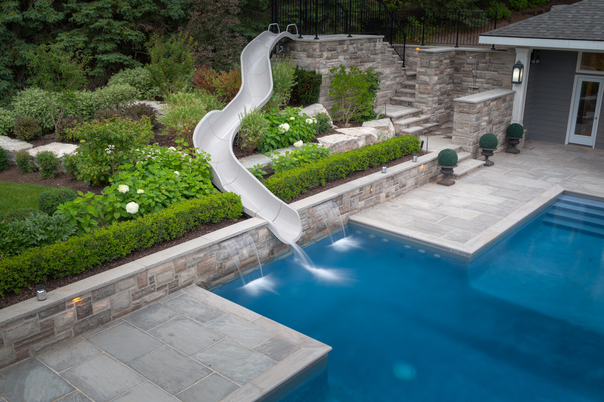 A backyard with an inground swimming pool and a twisting slide, surrounded by a landscaped garden and a stone patio on an overcast day.