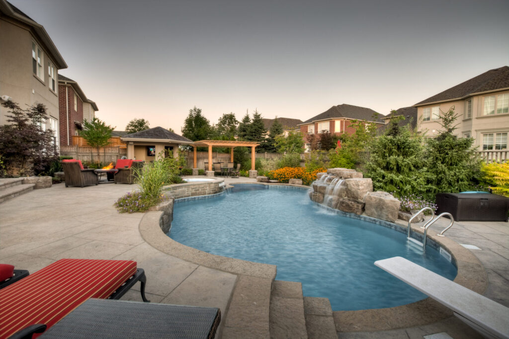 An inviting backyard with a curved pool, integrated hot tub, waterfall, stone patio, lounge chairs, gazebo, and lush landscaping during twilight.