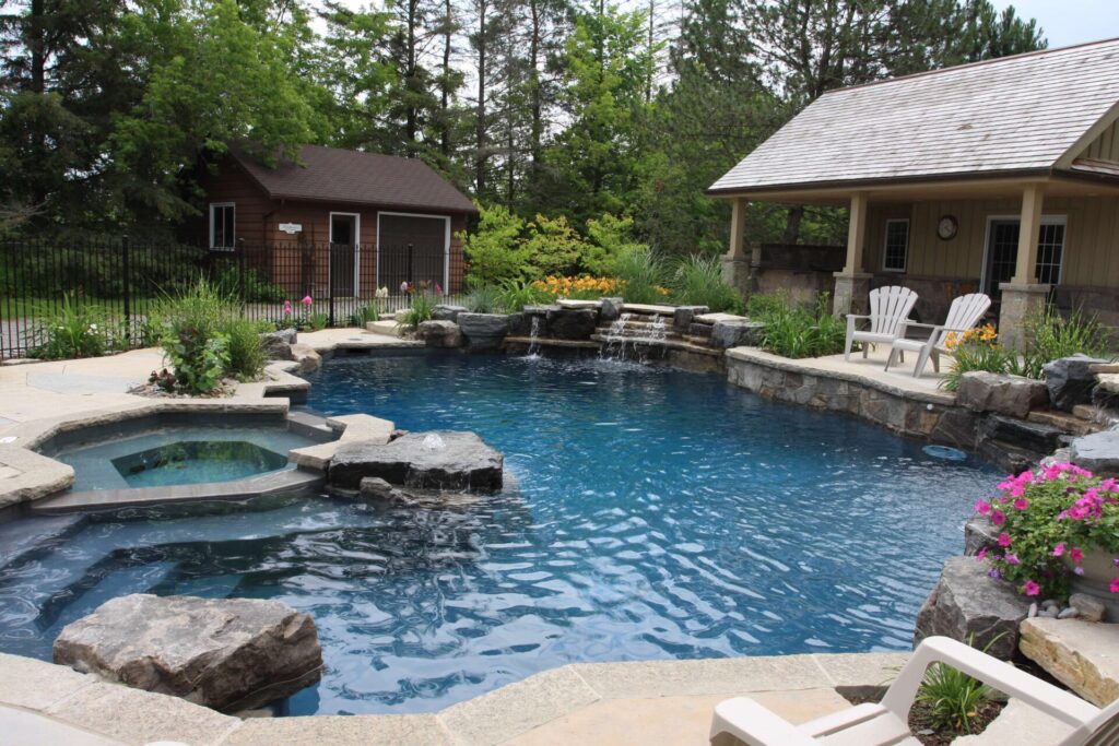 An inviting backyard with a landscaped pool featuring stone edges, a hot tub, greenery, a small outbuilding, lounger, and a garden chair.