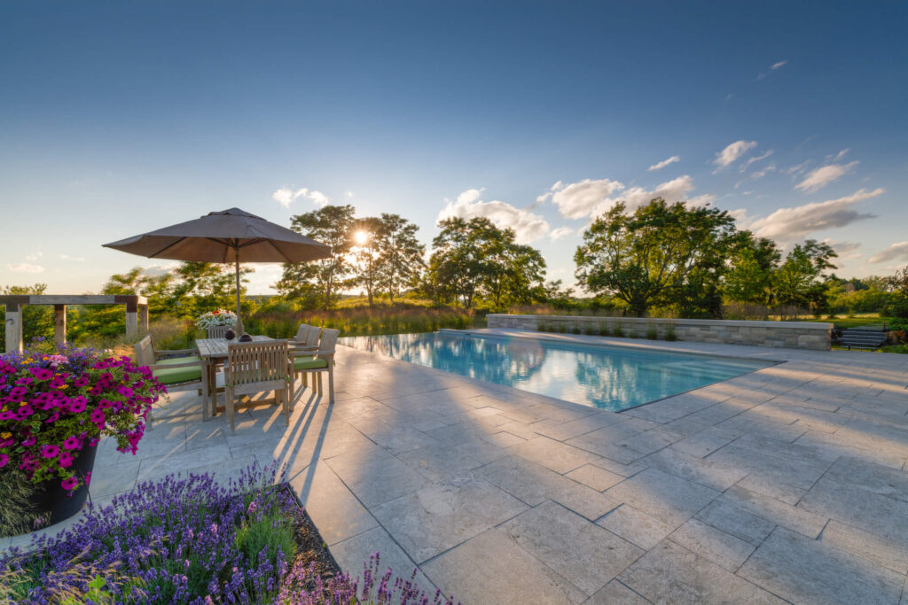 An outdoor patio with a swimming pool at sunset, featuring wooden furniture, an umbrella, flowering plants, and a scenic view with trees.