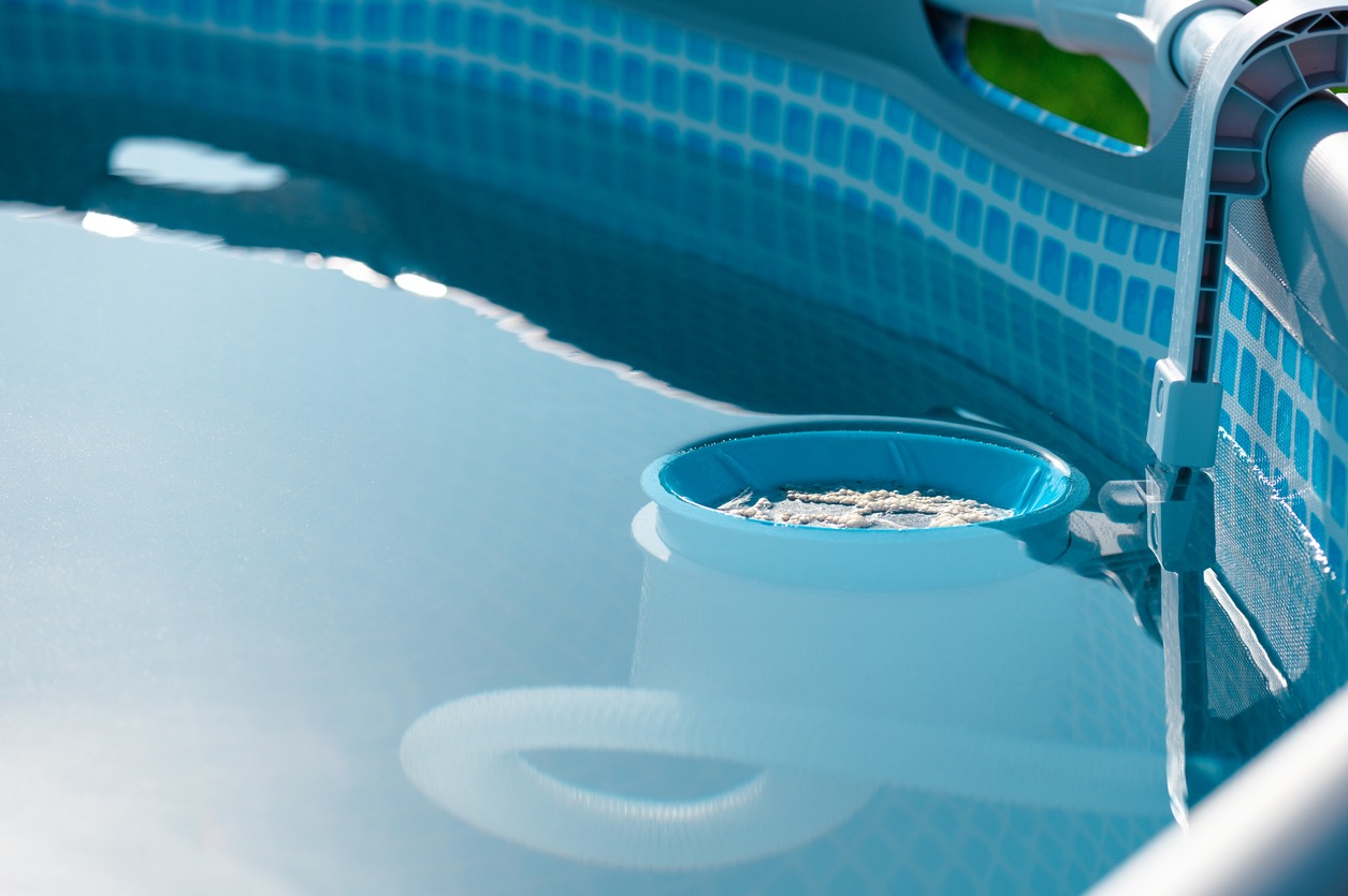 An above-ground pool with clear water, bordered by a blue mosaic pattern, with a pool skimmer floating on the surface under sunlight.