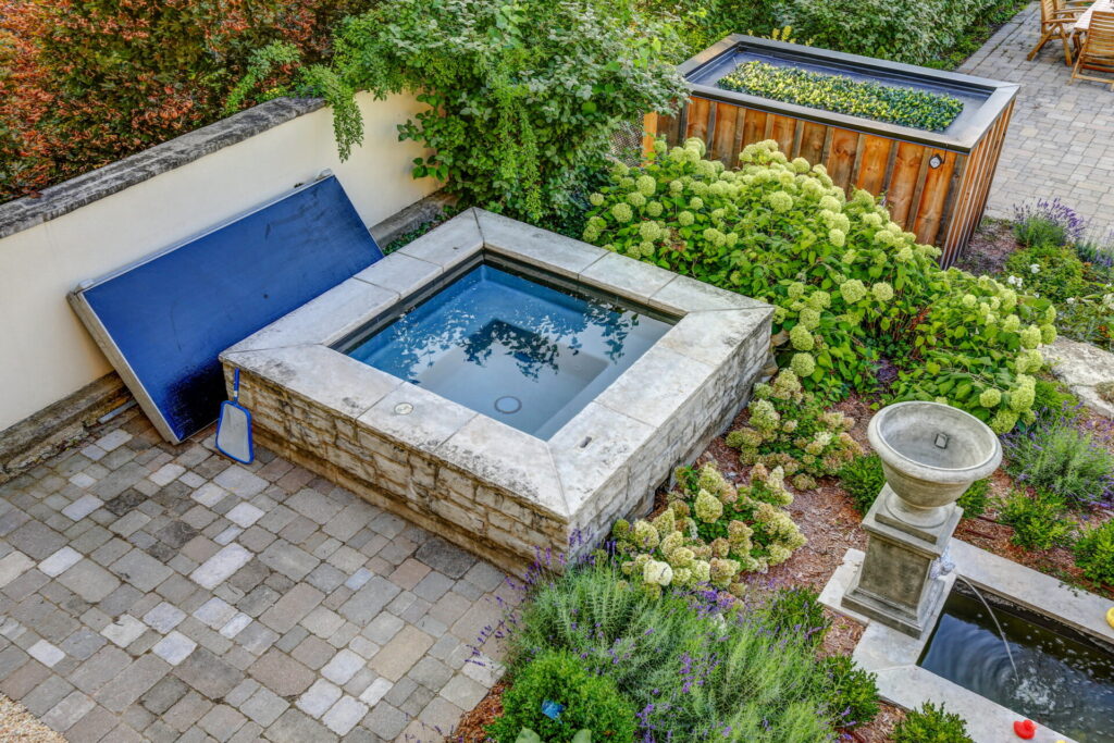 An elevated stone spa with a closed blue cover, amidst lush greenery, adjacent to a garden with a birdbath and wood-paneled planters.