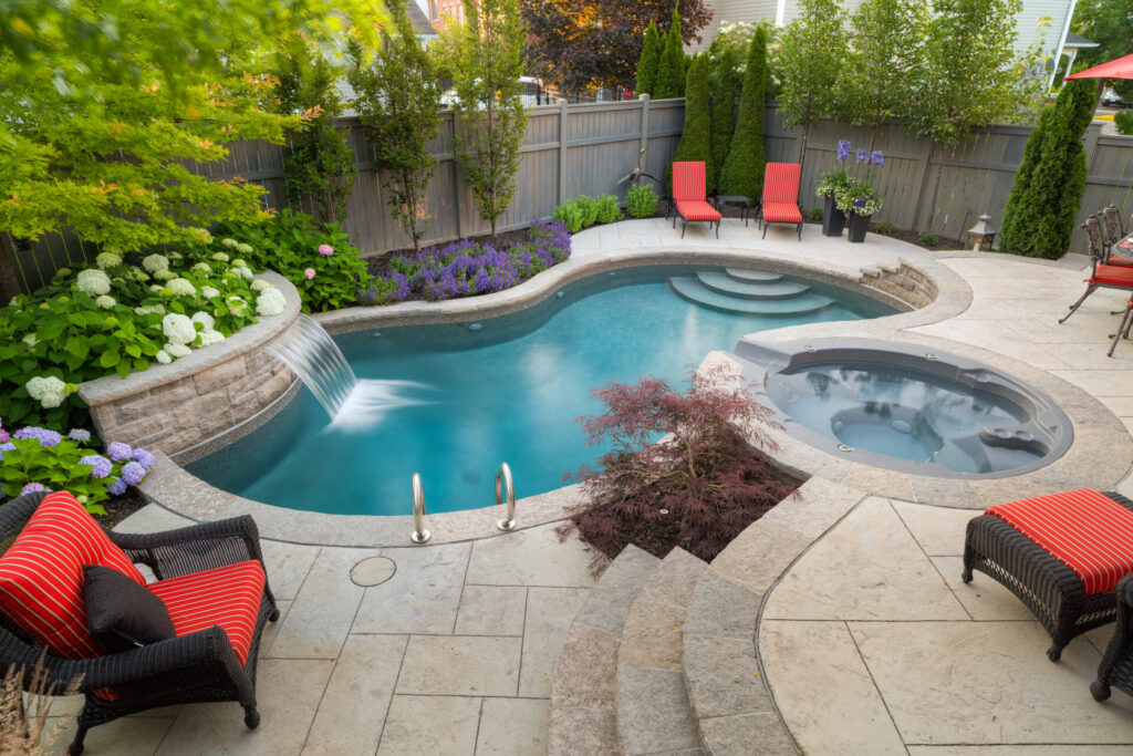 A luxurious backyard with a curved swimming pool, hot tub, waterfall, and vibrant landscaping. Outdoor furniture with red cushions adds a pop of color.