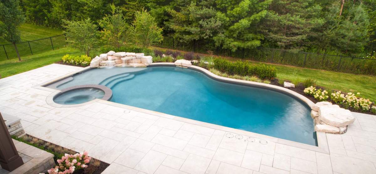 A kidney-shaped swimming pool with a jacuzzi, surrounded by a stone patio and lush landscaping, set against a backdrop of trees and grass.