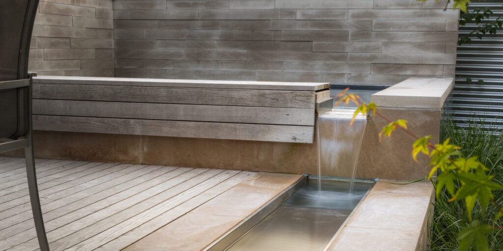 A modern outdoor space featuring a wooden bench, a flowing water feature, and subtle landscaping with plants, set against a textured grey wall.