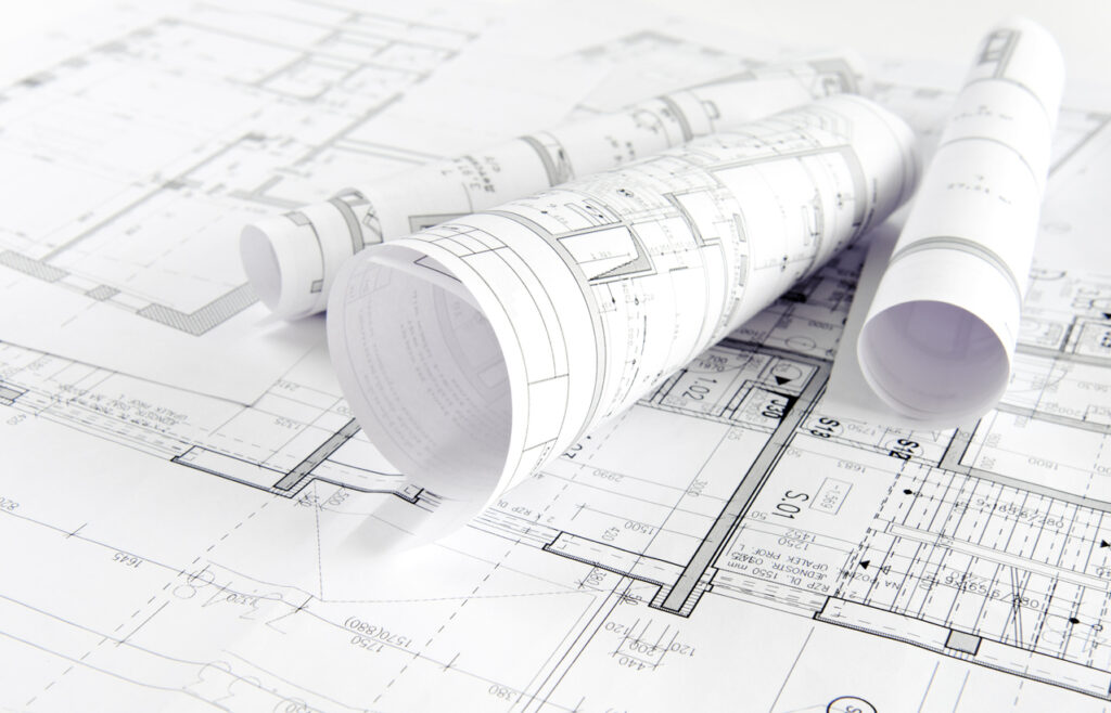The image shows rolled and unrolled architectural plans with detailed drawings for construction laid out on a white surface, indicating technical design work.