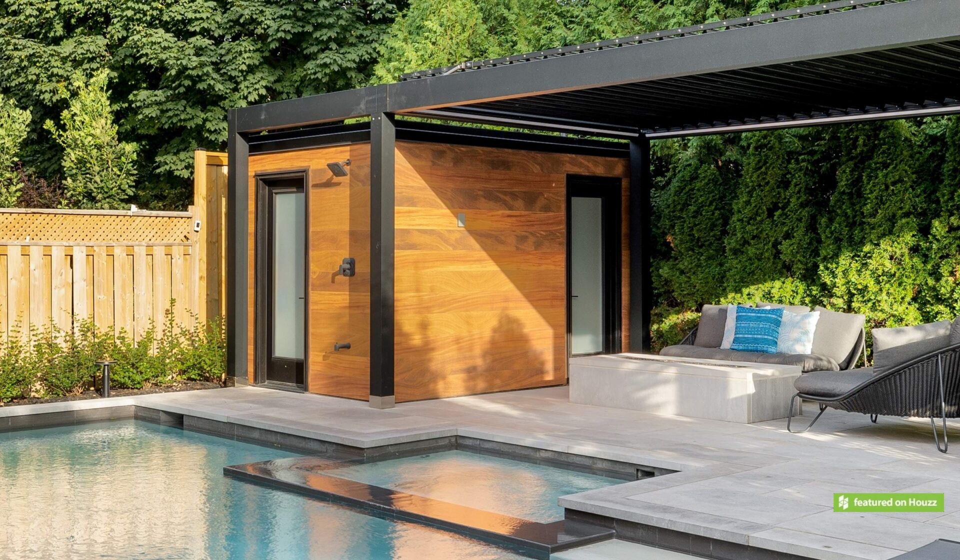 A modern poolside cabana with wooden walls and sliding door, under a pergola, next to an inground pool with a lounge chair and cushions.