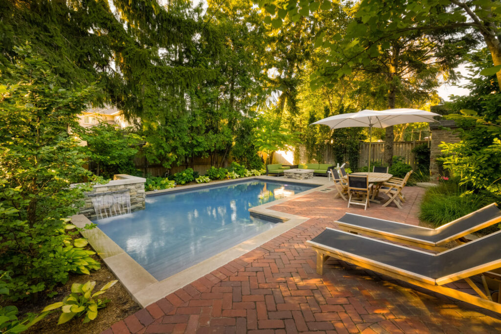 This image features an inviting backyard with a rectangular swimming pool, lounge chairs, an umbrella, outdoor furniture, surrounded by lush greenery and trees.