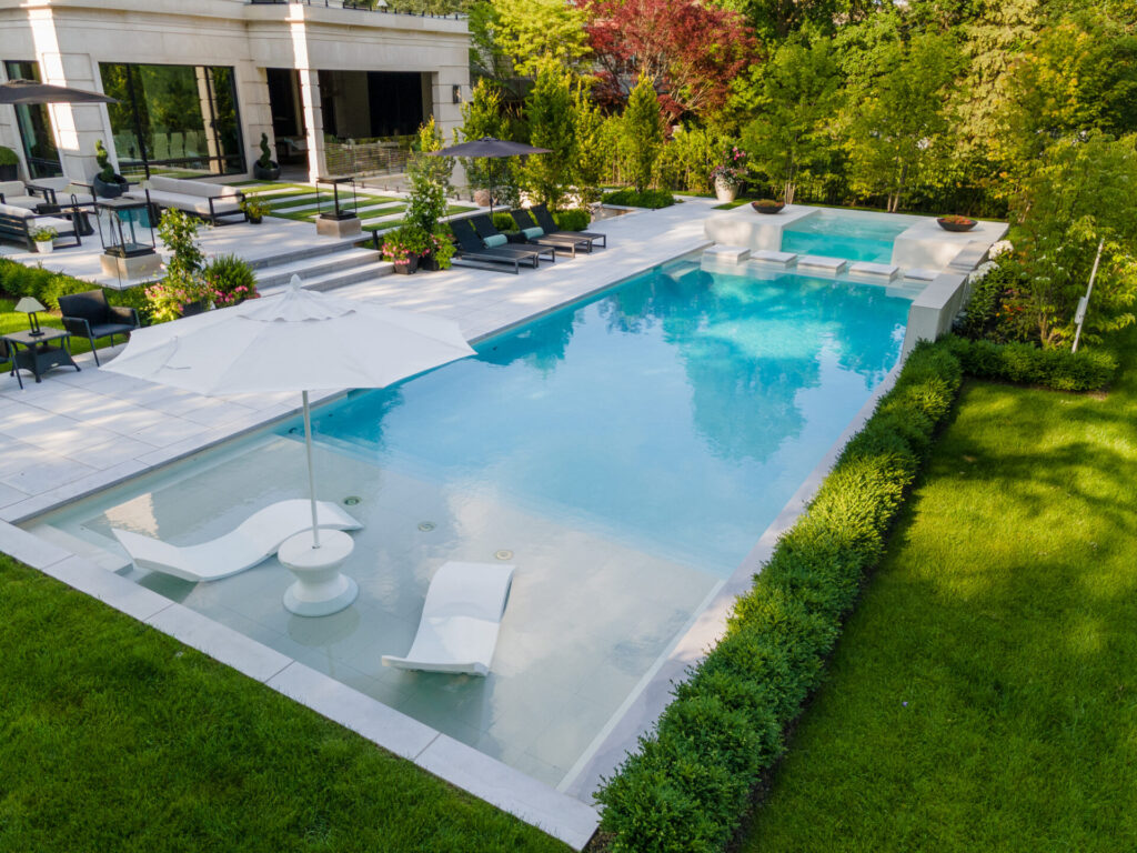 A luxurious backyard with a rectangular swimming pool, sun loungers, umbrellas, landscaped hedges, and an outdoor living space with modern furniture.