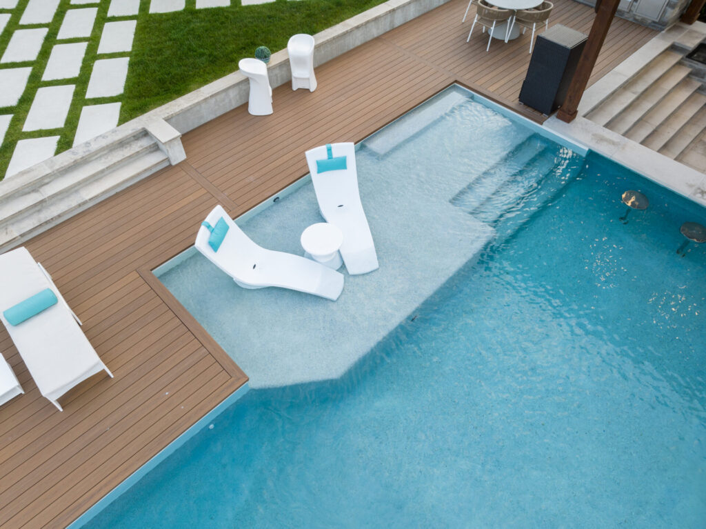 An aerial view of a residential backyard with a swimming pool featuring two white water slides, deck chairs, and a patterned lawn area.
