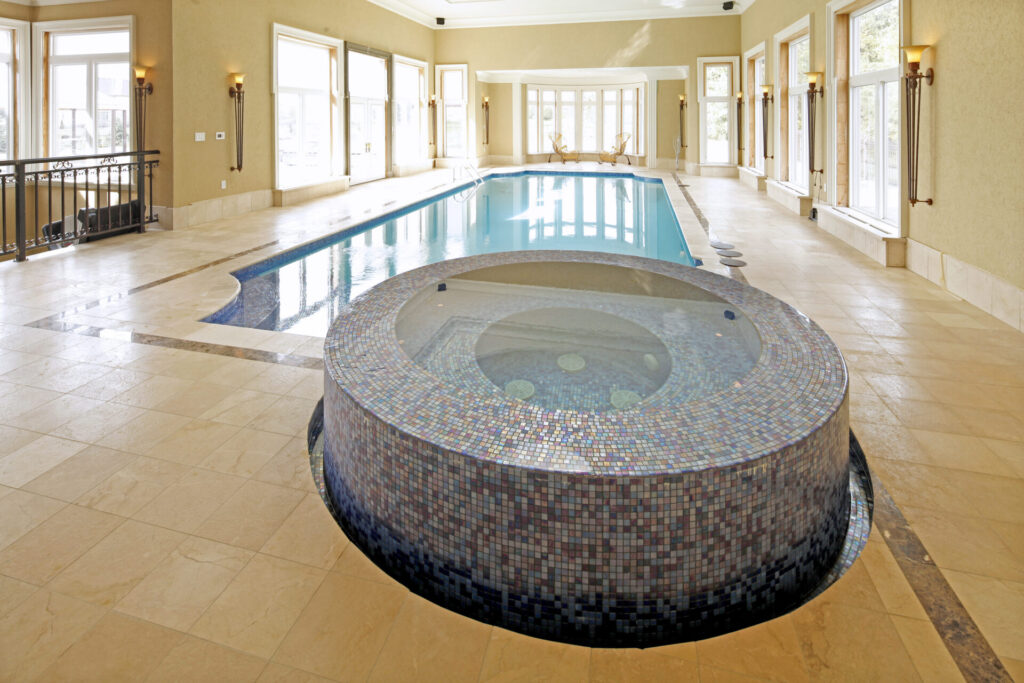 Indoor swimming pool with a hot tub in the center, surrounded by beige tiles, large windows, and multiple doors, under soft artificial lighting.