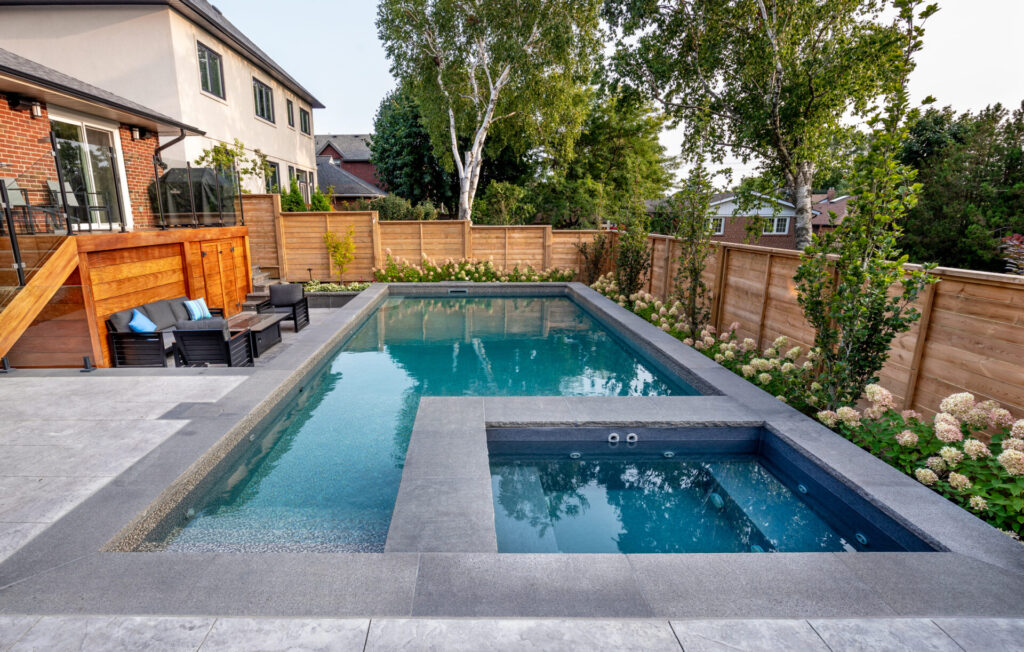 This image shows a modern backyard with a rectangular swimming pool, hot tub, wooden deck, outdoor furniture, landscaping, and a privacy fence.