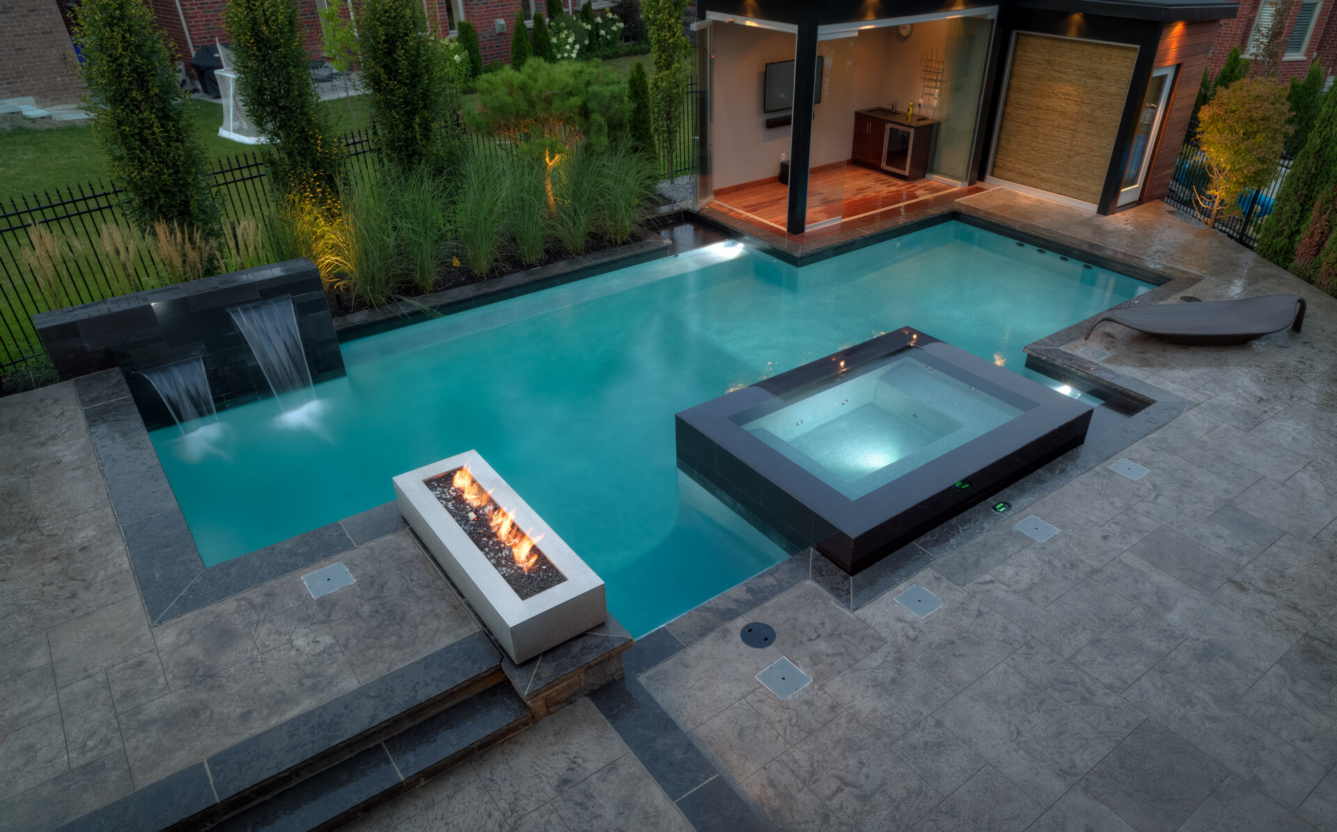 An elegant backyard featuring a swimming pool with a built-in jacuzzi and a waterfall, a fire pit, landscaped lighting, and an outdoor living space.