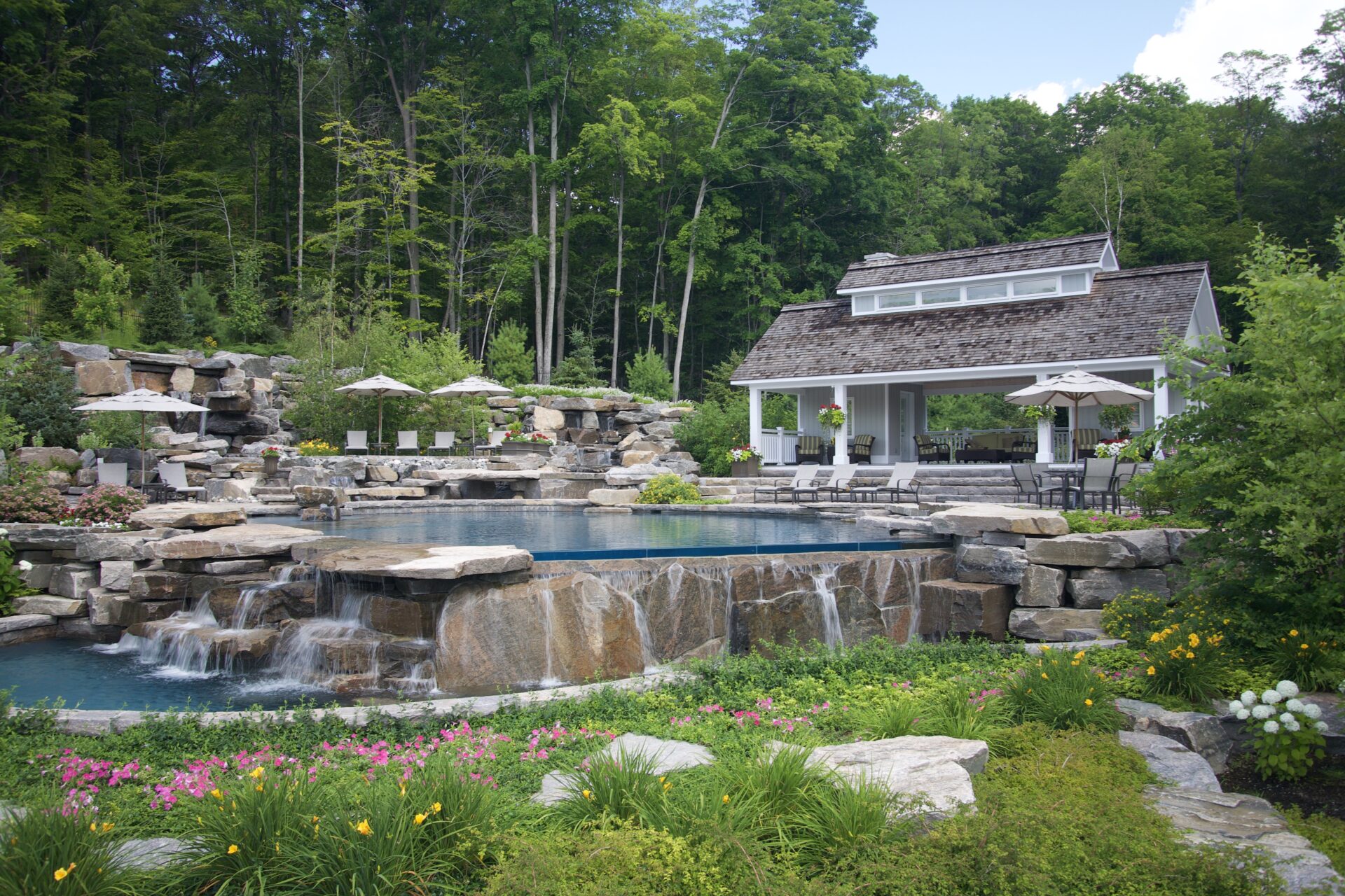 A luxurious backyard with an in-ground pool, waterfall, lush landscaping, patio area, and a pool house surrounded by trees under a clear sky.