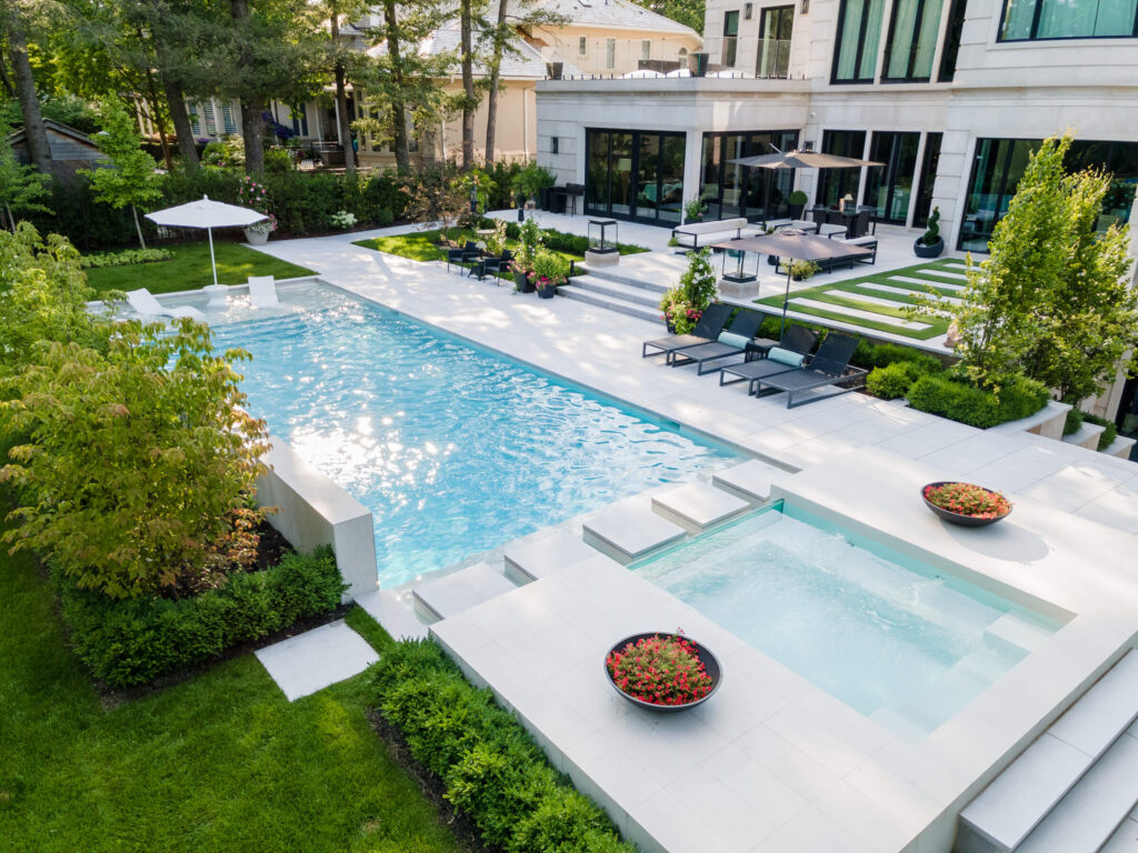 This image shows a luxurious backyard with a large swimming pool, a hot tub, well-manicured lawns, elegant outdoor furniture, and a modern house.