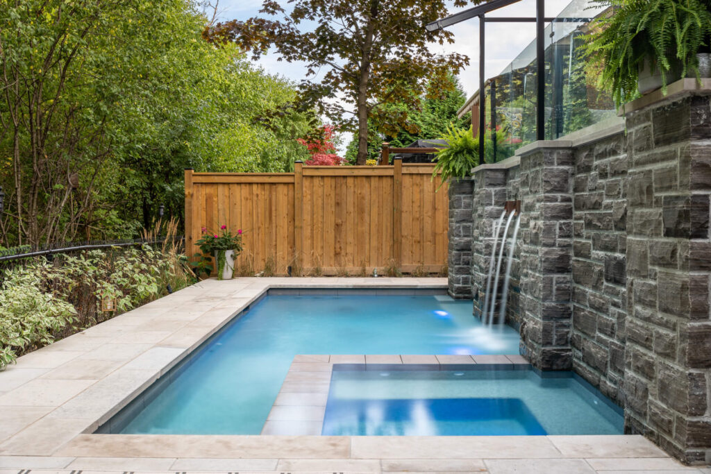 An outdoor swimming pool with a stone waterfall feature, surrounded by a wooden fence, lush plants, and a clear glass railing under cloudy skies.
