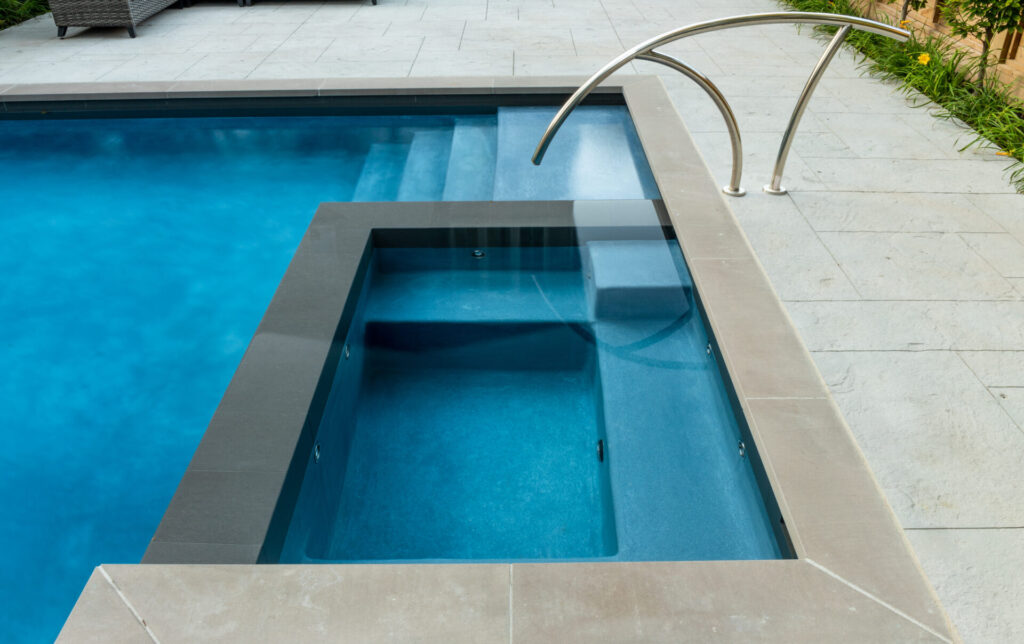 A modern swimming pool with clear blue water, featuring an adjacent smaller pool or hot tub and stainless steel handrails, surrounded by light paving stones.