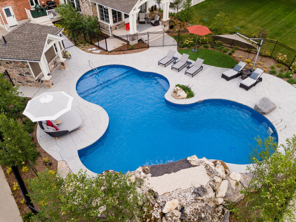 An aerial view of a backyard with a kidney-shaped swimming pool, stone patio, lounge chairs, umbrellas, landscaped garden, and a residential building.