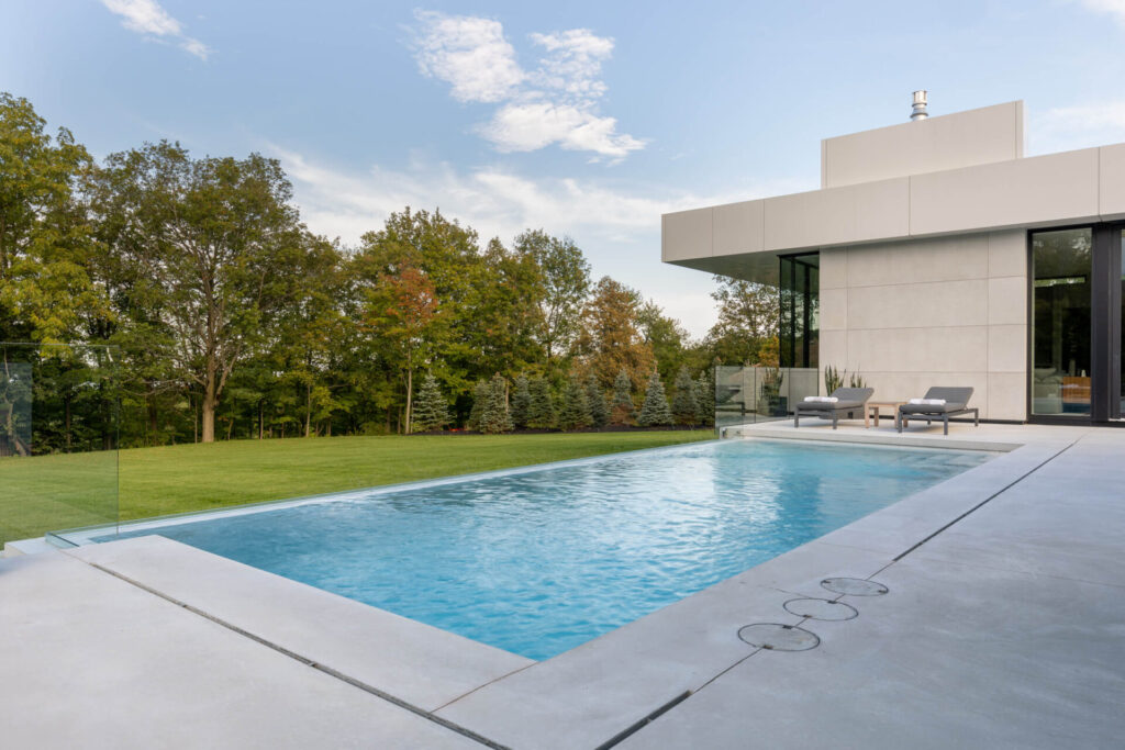 A serene outdoor setting with a swimming pool beside a modern house, lounge chairs, a well-kept lawn, and lush trees under a partly cloudy sky.