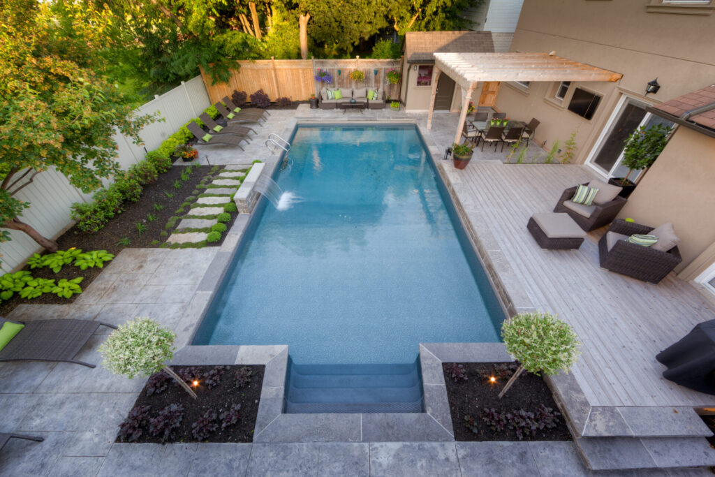 An aerial view of a backyard with a rectangular swimming pool, patio area with furniture, a pergola, and neatly landscaped garden beds.
