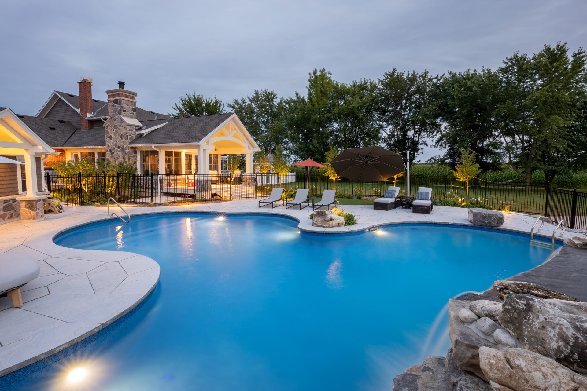 Luxurious backyard with an illuminated curved pool at dusk, surrounded by lounge chairs, landscaped garden, a cozy house, and a clear sky.