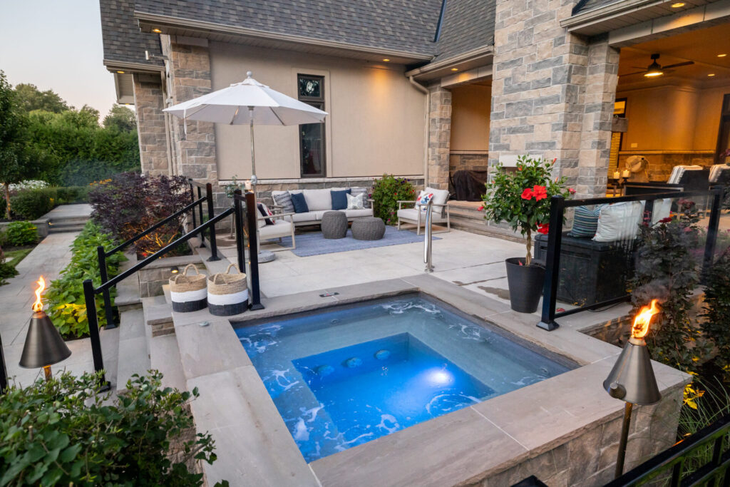 This image shows a modern outdoor patio with a small pool, comfortable seating area, umbrella, torches, plants, and a view into the interior.