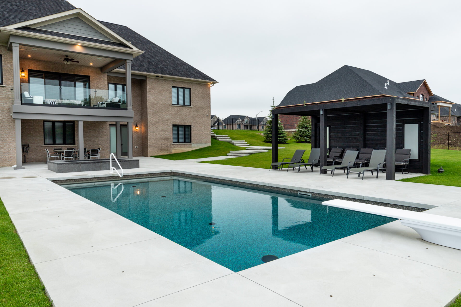 A modern two-story brick house with a balcony overlooks an outdoor rectangular swimming pool. A black pool house and patio chairs nearby complement the scene.