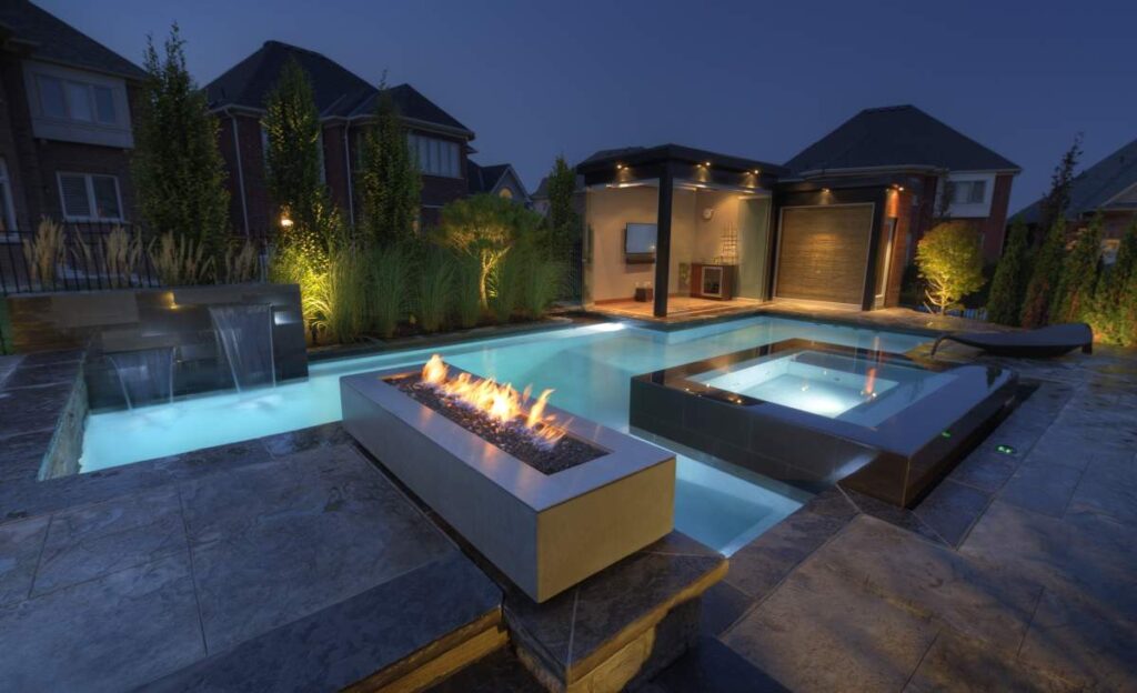 An outdoor evening setting includes a swimming pool with a water feature, a hot tub, a fire pit, lit landscaping, and a modern house background.