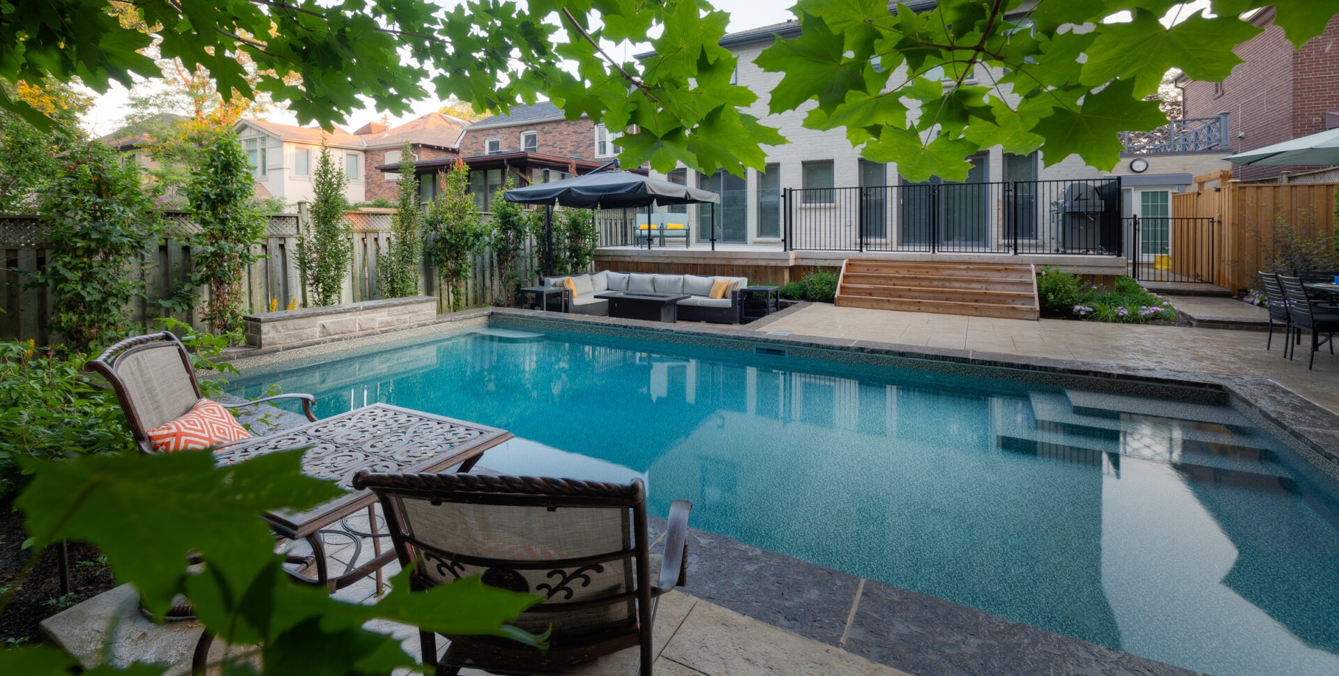 A tranquil backyard setting with a swimming pool, patio furniture, leafy trees, and surrounding residential buildings under a clear sky at dusk.